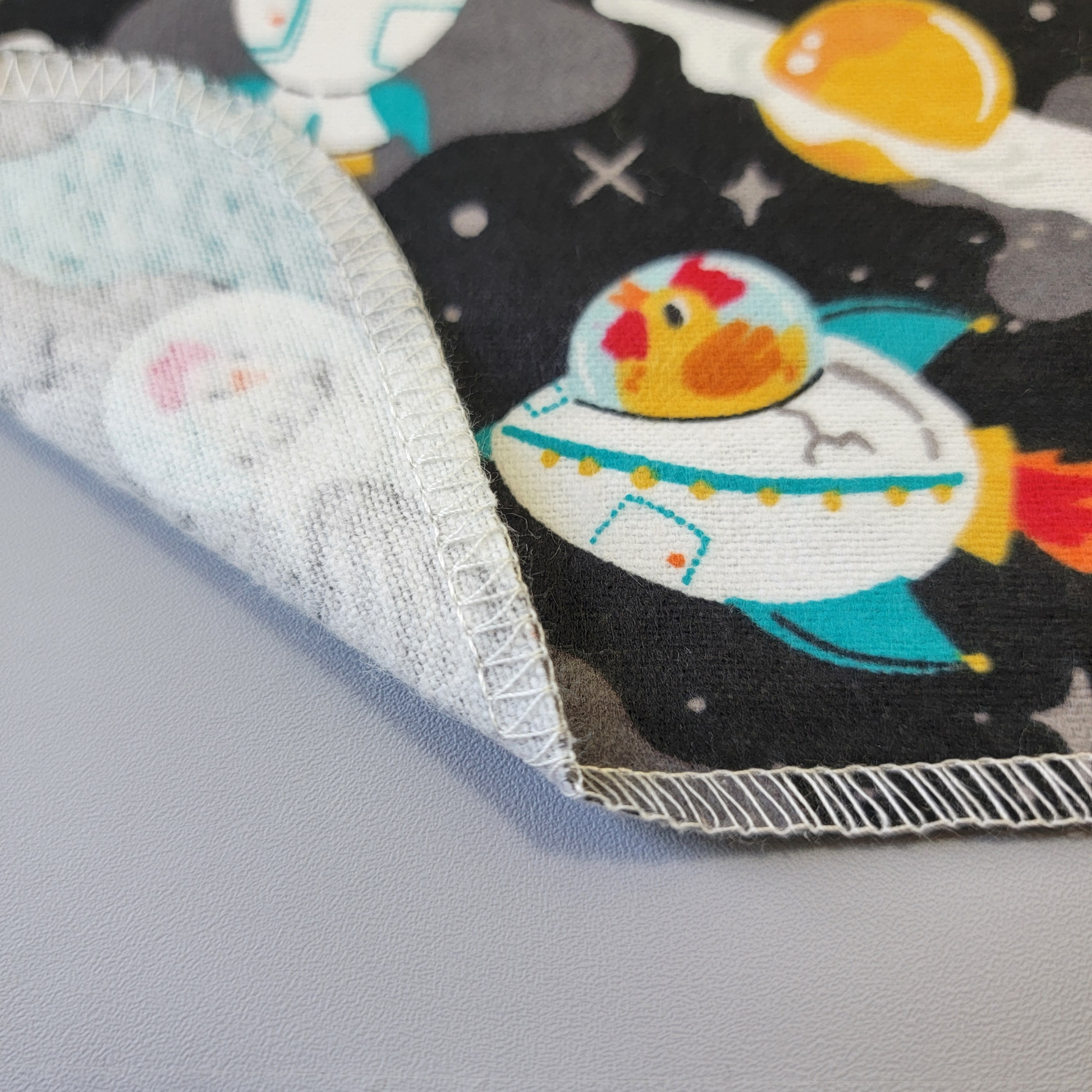 The bottom edge of a Space Chickens NonPaper Towel close-up to show the detail of the stitching. The edge is partially folded back to show the back side is unprinted, and there is one layer of material.