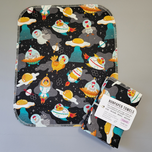 Space Chickens print NonPaper Towel laid flat to show the detail of the print, with a bundled 5-pack sitting partially on top of it. The NonPaper Towel is a rectangular shape, with rounded corners, The edges are stitched in off-white thread. The bundle's tag says "NonPaper Towels, the simplicity of a rag with the convenience of a paper towel. Use, wash, roll, reuse. 100% cotton fabric, 100% cellulose thread. Plant based, biodegradable."