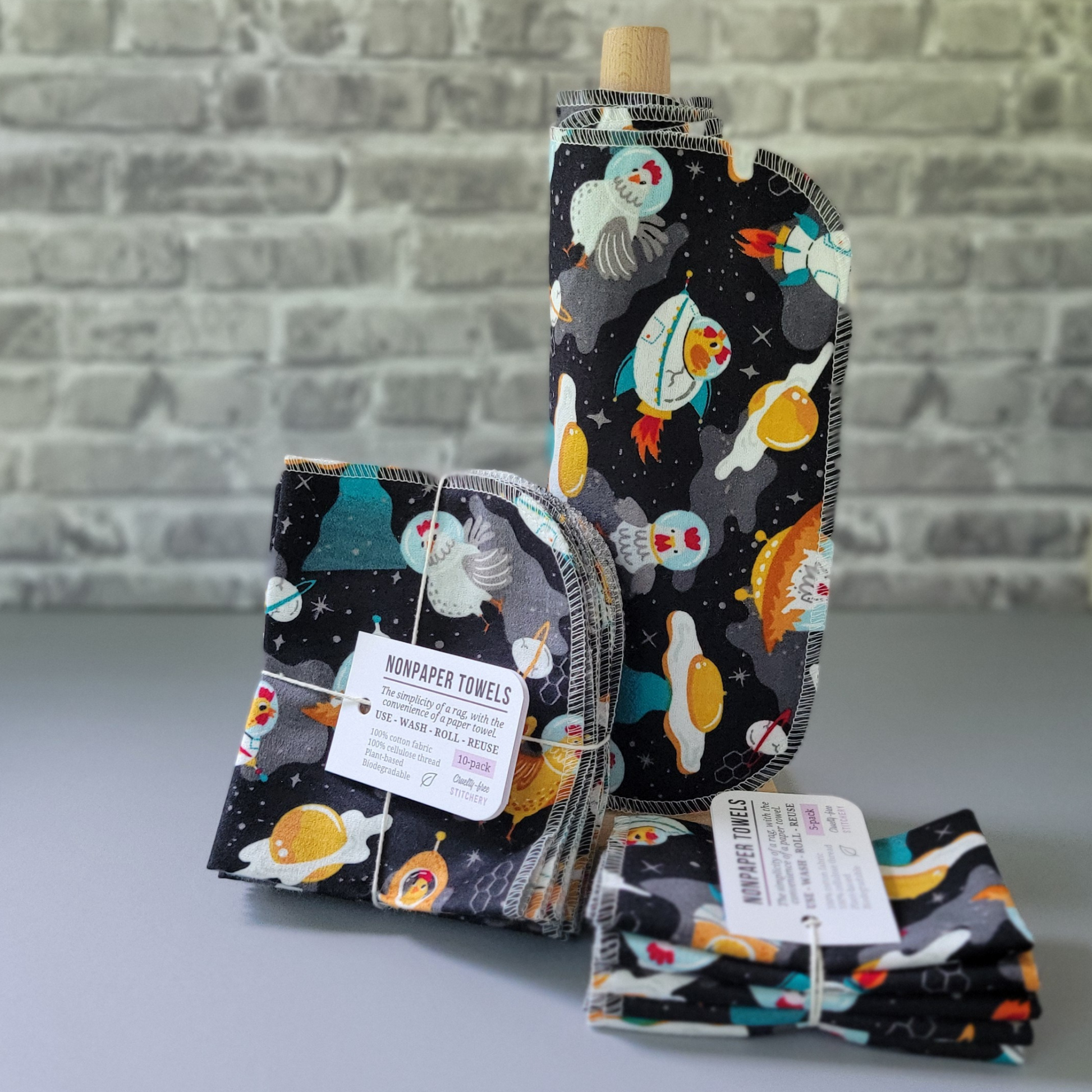 Space Chickens print NonPaper Towels. The print is cartoonish chickens in space, some in space helmets, some in rockets and flying saucers. The planets are eggs. In the center, they are rolled onto a wooden paper towel holder, and there are bundled packs of 10 and 5 sitting next to the holder. The bundles are tied with string and have a small paper tag. The background is a gray brick wall and solid gray tabletop.