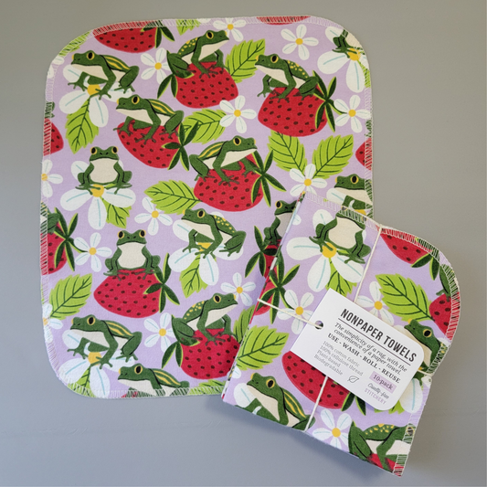 Strawberry Frogs print NonPaper Towel laid flat to show the detail of the print, with a bundled 10-pack sitting partially on top of it. The NonPaper Towel is a rectangular shape, with rounded corners, The edges are stitched in off-white thread. The bundle's tag says "NonPaper Towels, the simplicity of a rag with the convenience of a paper towel. Use, wash, roll, reuse. 100% cotton fabric, 100% cellulose thread. Plant based, biodegradable."