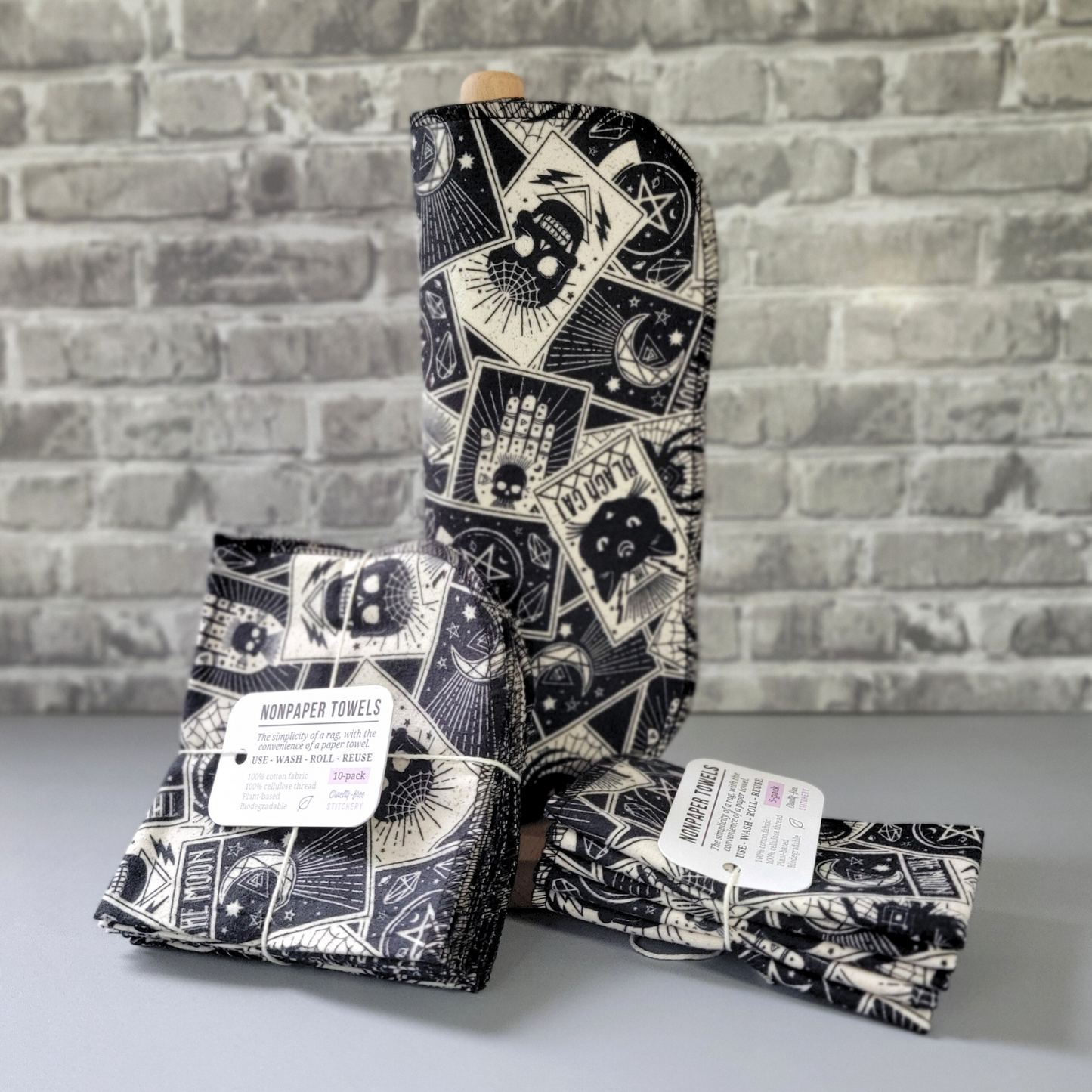 Tarot cards print nonpaper towels, displayed on a wooden paper towel holder, laid next to bundled 5 and 10-packs. The towels are black and off-white, with a print that looks like scattered tarot cards. Black cat, the moon, spider, palms, pentacles, etc.