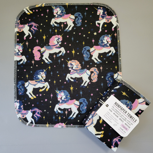 Unicorns print NonPaper Towel laid flat to show the detail of the print, with a bundled 5-pack sitting partially on top of it. The NonPaper Towel is a rectangular shape, with rounded corners, The edges are stitched in off-white thread. The bundle's tag says "NonPaper Towels, the simplicity of a rag with the convenience of a paper towel. Use, wash, roll, reuse. 100% cotton fabric, 100% cellulose thread. Plant based, biodegradable."