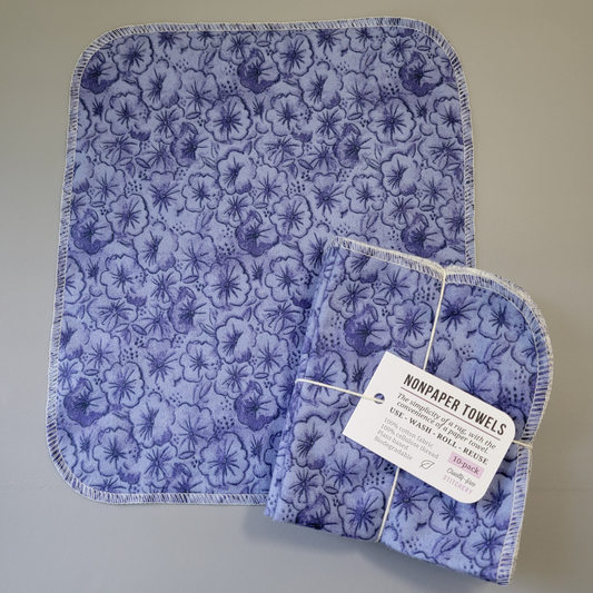 Violets print NonPaper Towel laid flat to show the detail of the print, with a bundled 10-pack sitting partially on top of it. The NonPaper Towel is a rectangular shape, with rounded corners, The edges are stitched in off-white thread. The bundle's tag says "NonPaper Towels, the simplicity of a rag with the convenience of a paper towel. Use, wash, roll, reuse. 100% cotton fabric, 100% cellulose thread. Plant based, biodegradable."