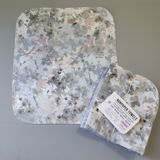Watercolor Floral Gray print NonPaper Towel laid flat to show the detail of the print, with a bundled 10-pack sitting partially on top of it. The NonPaper Towel is a rectangular shape, with rounded corners, The edges are stitched in off-white thread. The bundle's tag says "NonPaper Towels, the simplicity of a rag with the convenience of a paper towel. Use, wash, roll, reuse. 100% cotton fabric, 100% cellulose thread. Plant based, biodegradable."