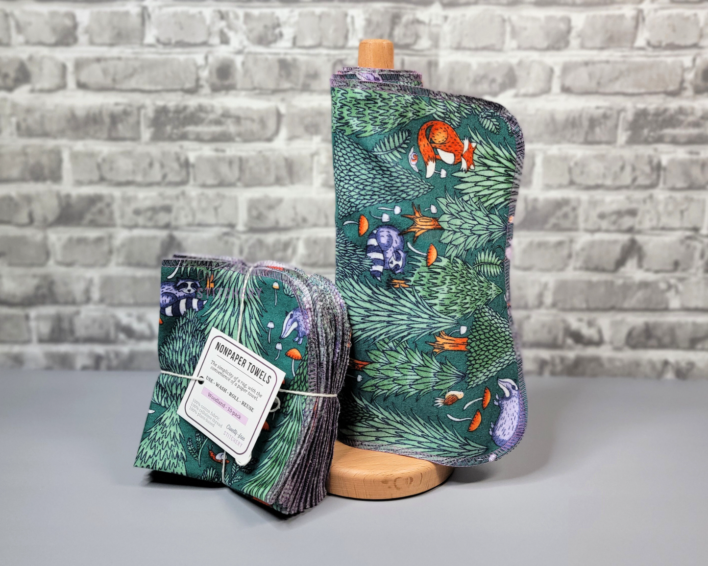 Woodland print NonPaper Towels. Displayed on a wooden paper towel holder next to a bundled pack. They are dark green with trees, mushrooms, and various animals like foxes, raccoons, etc.