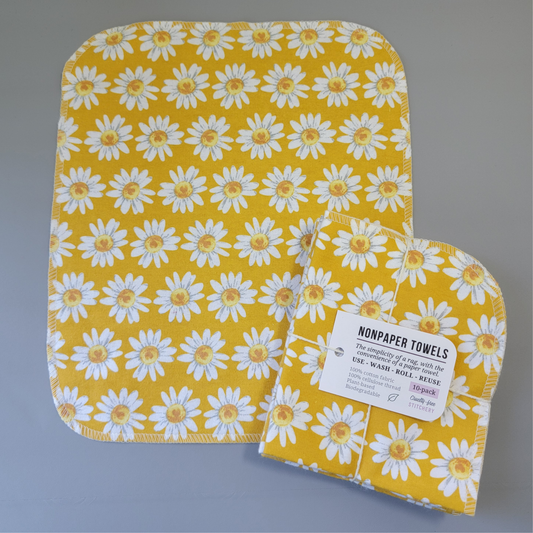 Yellow daisies print NonPaper Towel laid flat to show the detail of the print, with a bundled 10-pack sitting partially on top of it. The NonPaper Towel is a rectangular shape, with rounded corners, The edges are stitched in off-white thread. The bundle's tag says "NonPaper Towels, the simplicity of a rag with the convenience of a paper towel. Use, wash, roll, reuse. 100% cotton fabric, 100% cellulose thread. Plant based, biodegradable."