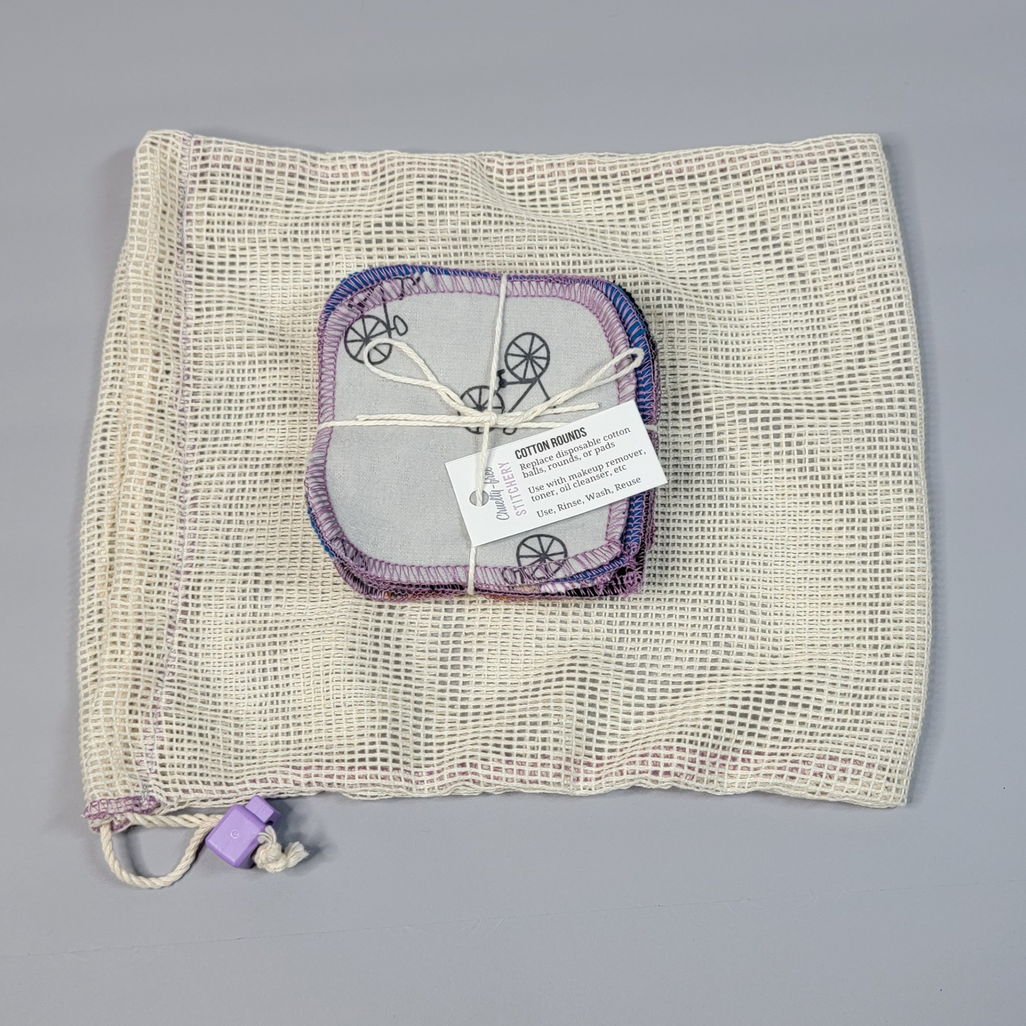 Bundled pack of cotton rounds on top of a mesh drawstring bag. The bag is a natural off-white colored square mesh, with a light purple cord stop.