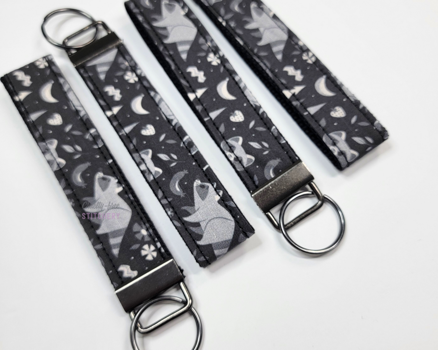Wristlet key fobs arranged in a row of four with ends alternating. The wristlet is black fabric printed with grey raccoons, trees, and food. The hardware and key ring are gunmetal silver.