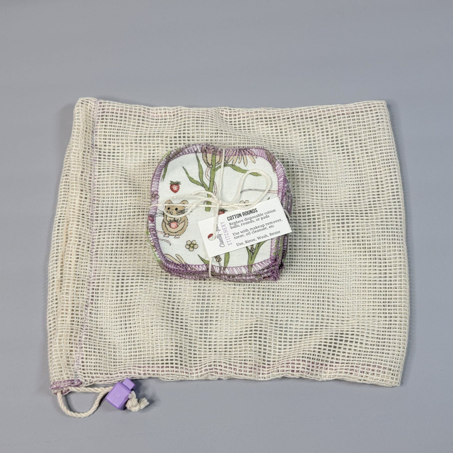 Bundled pack of cotton rounds on top of a mesh drawstring bag. The bag is a natural off-white colored square mesh, with a light purple cord stop.