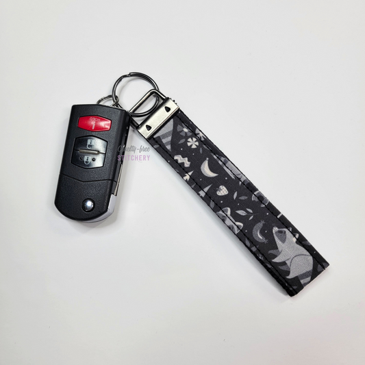 Raccoons print key fob wristlet attached to a car key for scale. The car key is the fold-away type and is about half as long as the strap.