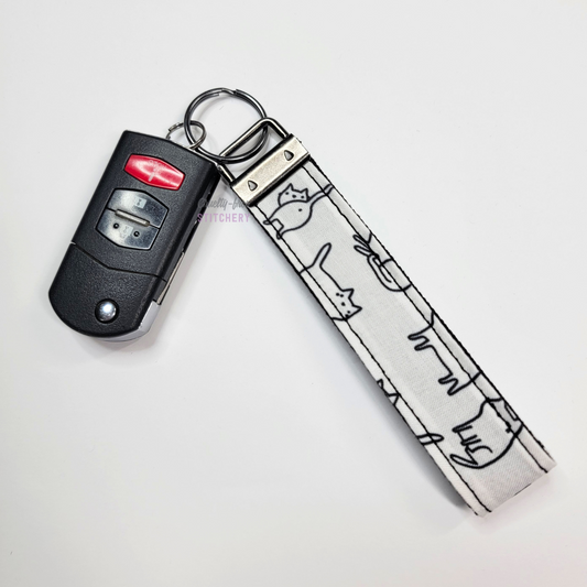 White cats print key fob wristlet attached to a car key for scale. The car key is the fold-away type and is about half as long as the strap.