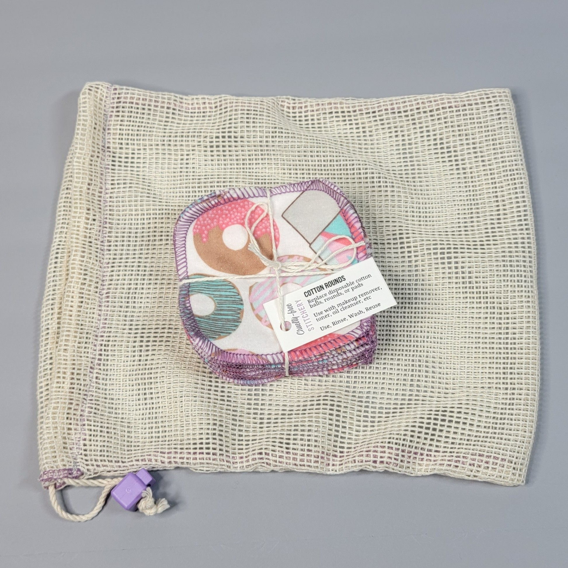 Bundled pack of cotton rounds on top of a mesh drawstring bag. The bag is a natural off-white colored square mesh, with a light purple cord stop.