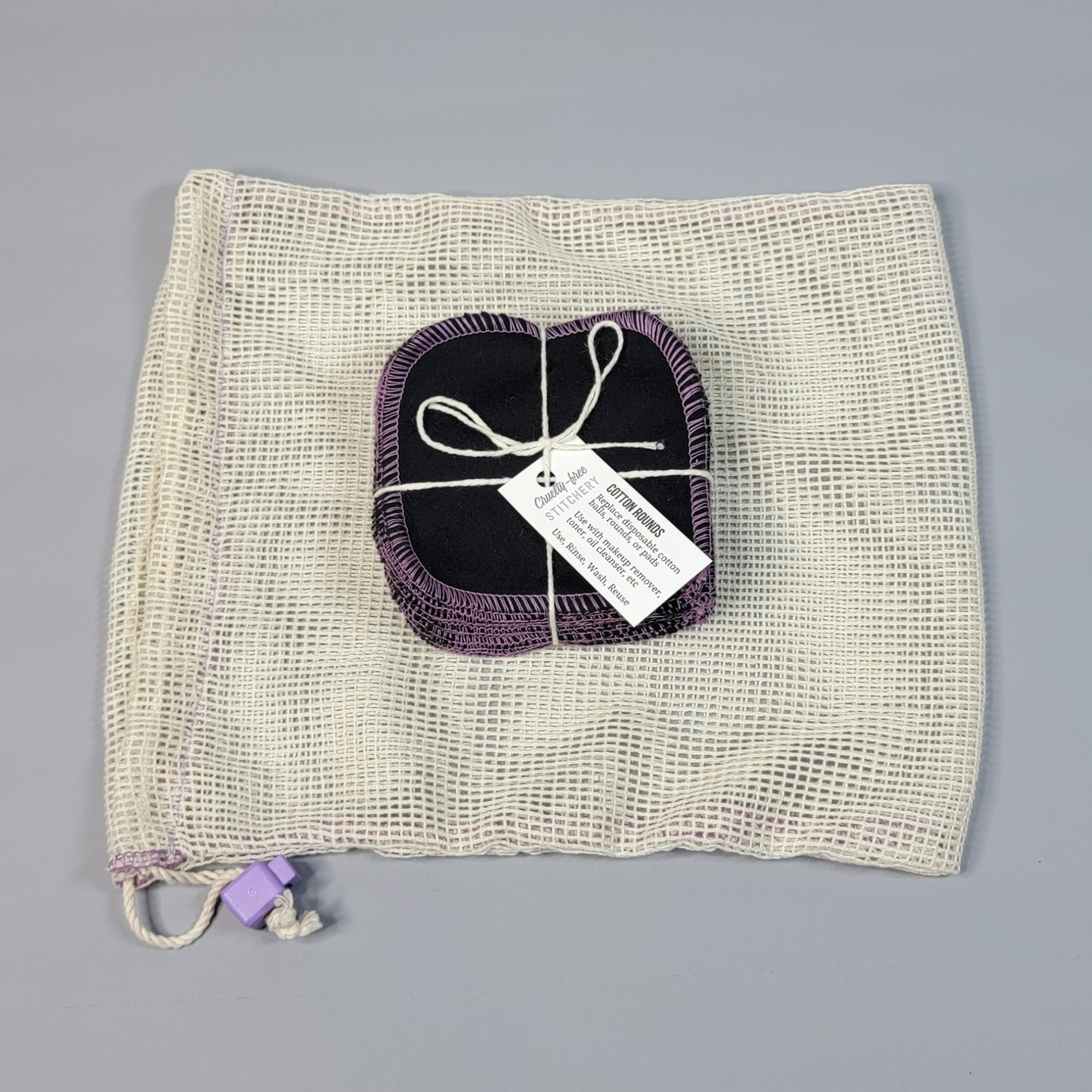 Bundled pack of cotton rounds on top of a mesh drawstring bag. The bag is a natural off-white colored square mesh, with a light purple cord stop.