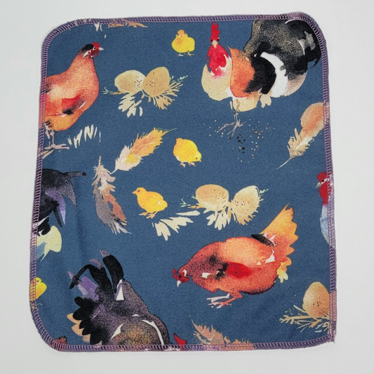 a blue chickens NonPaper Towel laid flat to show the print and size. There are a few farmhouse style chickens, about 4 to 6 inches wide, and some small one inch eggs, chicks, and feathers scattered around. The edges are rounded and stitched with a lilac purple thread.