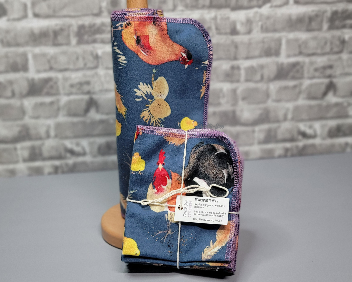 A set of deep blue NonPaper Towels with large chickens, eggs, and little yellow chicks in a realistic painted style. One set is rolled around a wooden holder, the other is folded into a bundle and sitting in front of the holder.