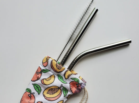 Reusable straw pouch with stainless steel straws sticking out the top. The fabric of the pouch is white with cartoon peaches printed on, some whole peaches and others are half or in slices. The pouch has a drawstring closure.