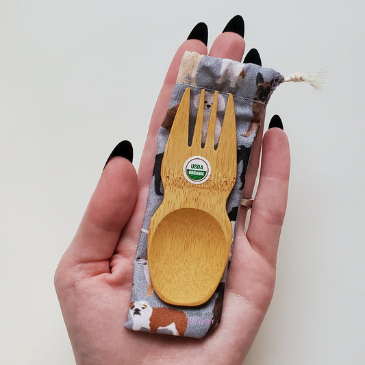 Reusable bamboo spork with pouch. Dog print fabric pouch with bamboo spork on top, laid on a hand for size reference. The spork is slightly longer than the fingers.