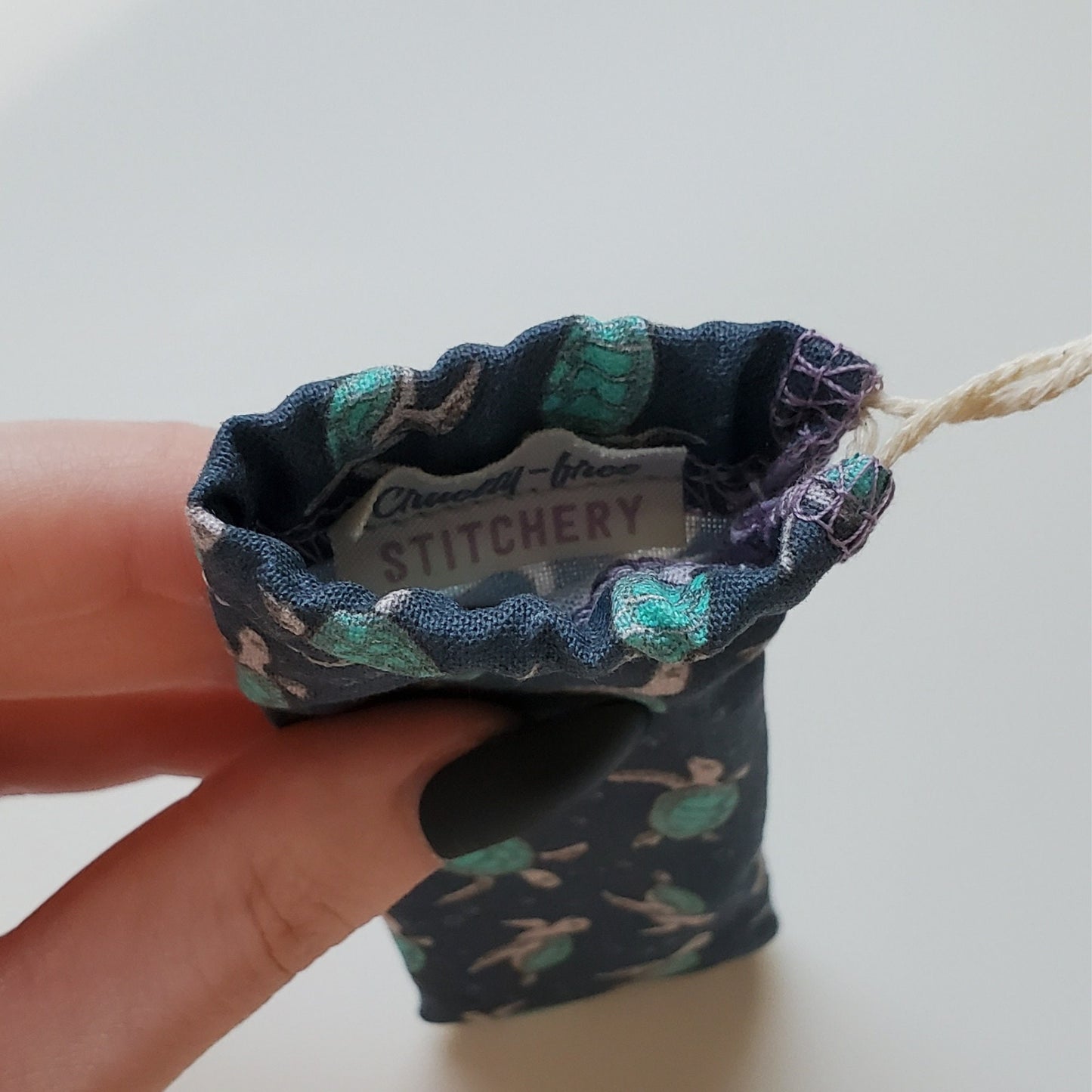 A reusable spork pouch, viewed from the top down. Inside the pouch is a tag with the Cruelty-Free Stitchery logo. The pouch is in someone's hand, the width is about half as wide as the thumb is long.