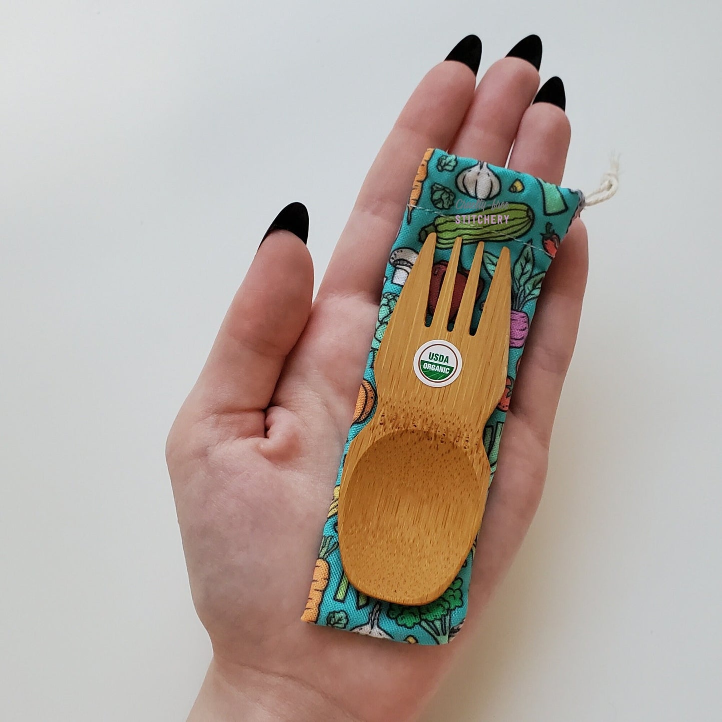 Reusable bamboo spork with pouch. Vegetable print fabric pouch with bamboo spork on top, laid on a hand for size reference. The spork is slightly longer than the fingers.