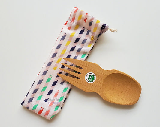 Reusable bamboo spork with pouch. The pouch is white fabric with multicolor  paper straws printed on. The pouch is sitting diagonally with the spork partially on top pointing the other way. The spork is small, a double ended fork and spoon.
