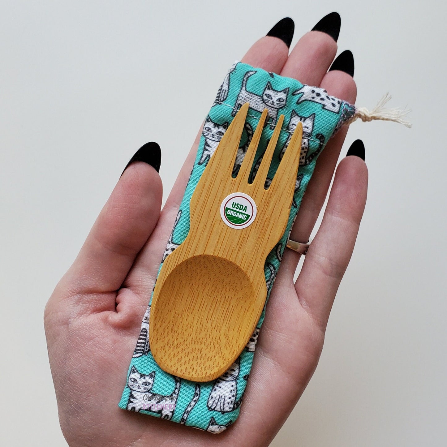 Reusable bamboo spork with pouch. Cat print fabric pouch with bamboo spork on top, laid on a hand for size reference. The spork is slightly longer than the fingers.
