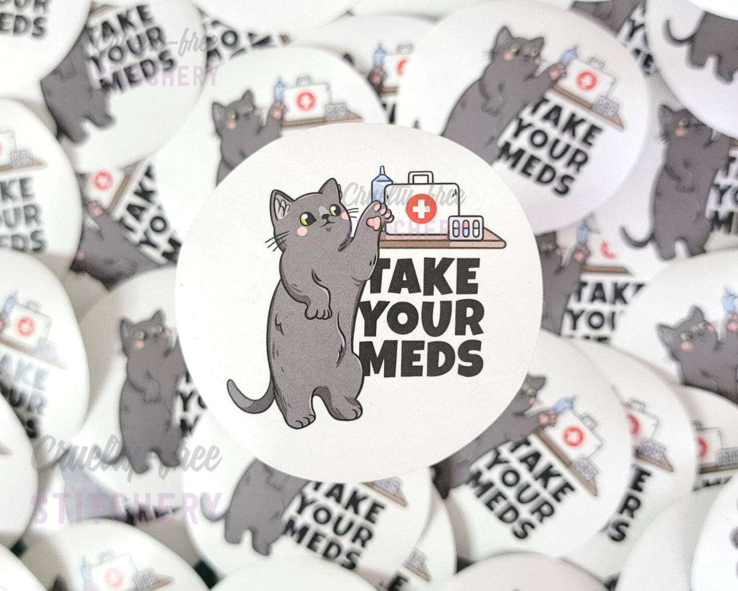 Round sticker that says "take your meds" with a cartoon grey cat reaching for a shelf with a first-aid kit and medicines.