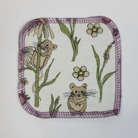 The front of one mouse print reusable cotton round. The fabric is off-white, with small light brown mice, green plants, red strawberries, and dandelions. One mouse is climbing a dandelion.