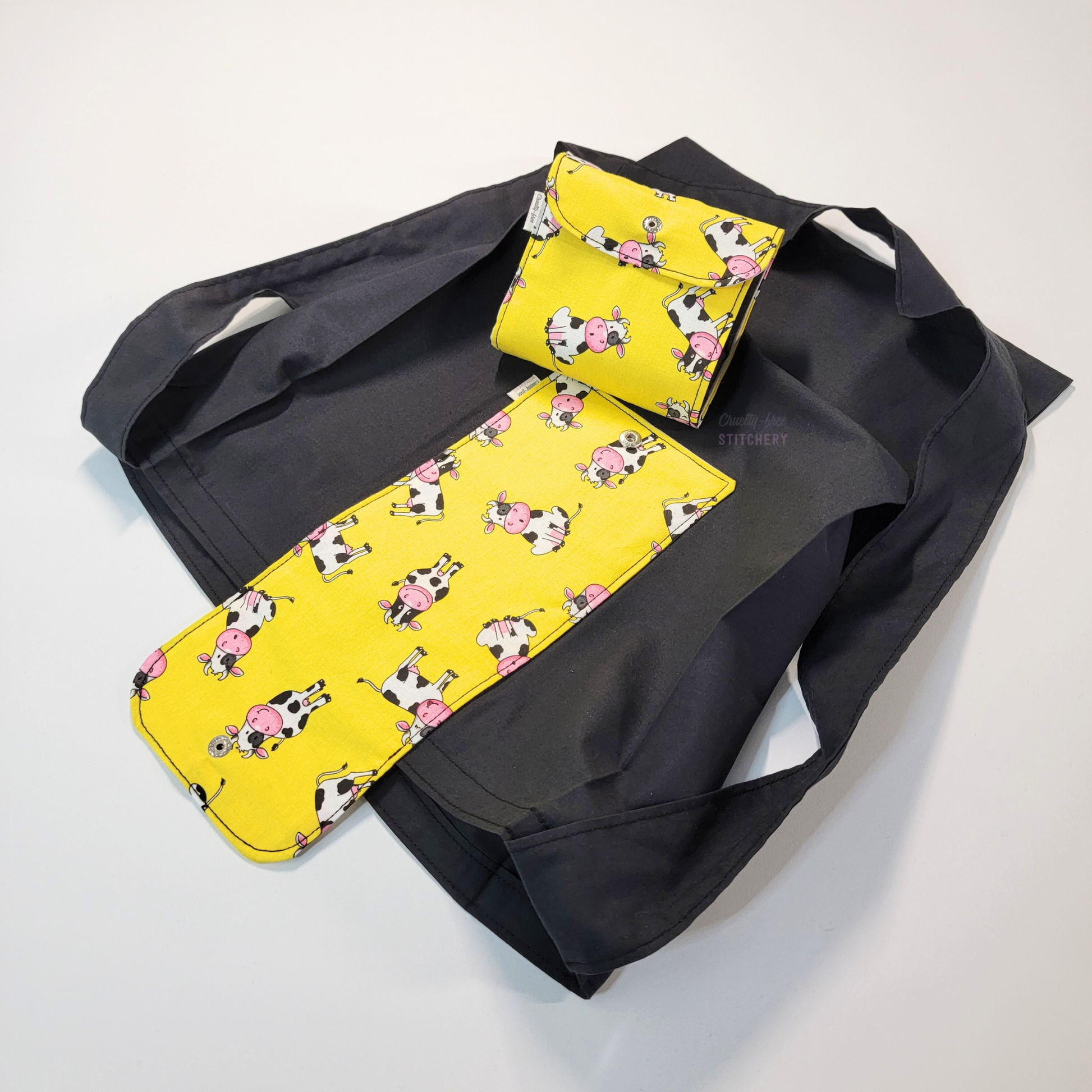 A solid black tote bag with a long strap and a bright yellow flap printed with cute smiling sketched cows in various poses. On top of the bag is another bag that has been folded up and snapped together like a wallet.