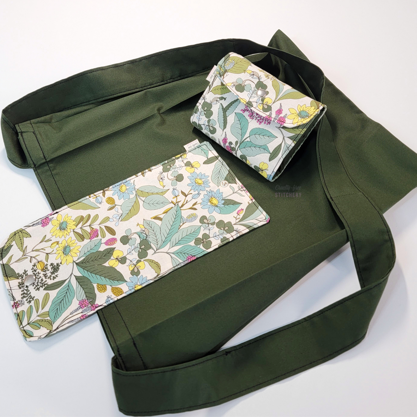 A solid dark forest green tote bag with a long strap and a light tan, sketched plants print flap. On top of the bag is another bag that has been folded up and snapped together like a wallet.