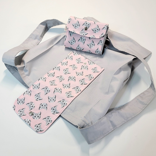 A solid pale gray tote bag with a long strap and a light pink flap printed with small greyscale tabby cat heads. On top of the bag is another bag that has been folded up and snapped together like a wallet.