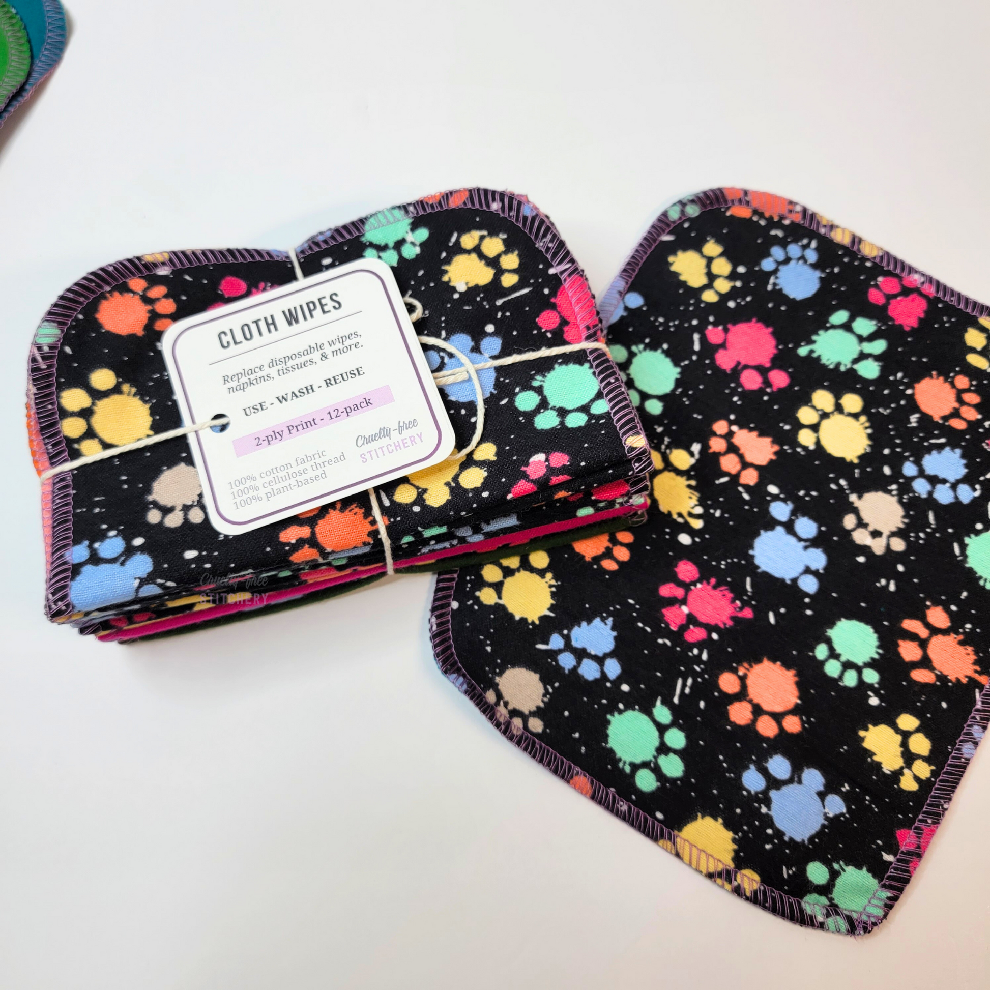 A bundled pack of the rainbow paw print paint splatter cloth wipes, next to an extra one laid flat. The print on the front is a black background with the paw print paint splatter design in bright colors.