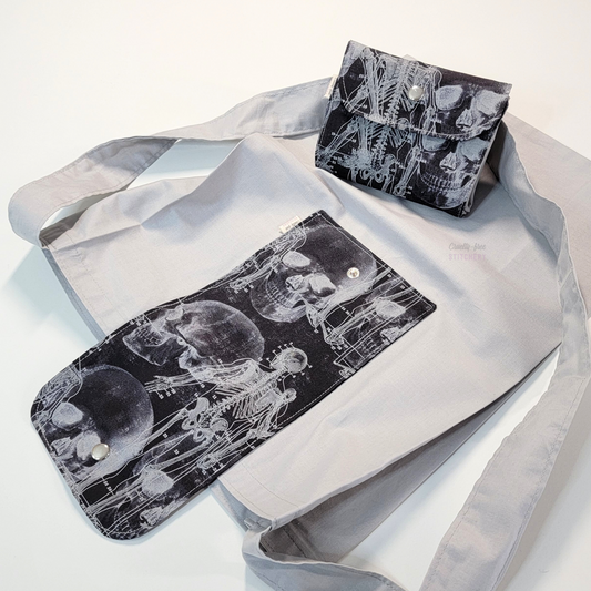 A solid pale gray tote bag with a long strap and a black flap printed with x-ray images of a skull and skeleton. On top of the bag is another bag that has been folded up and snapped together like a wallet.