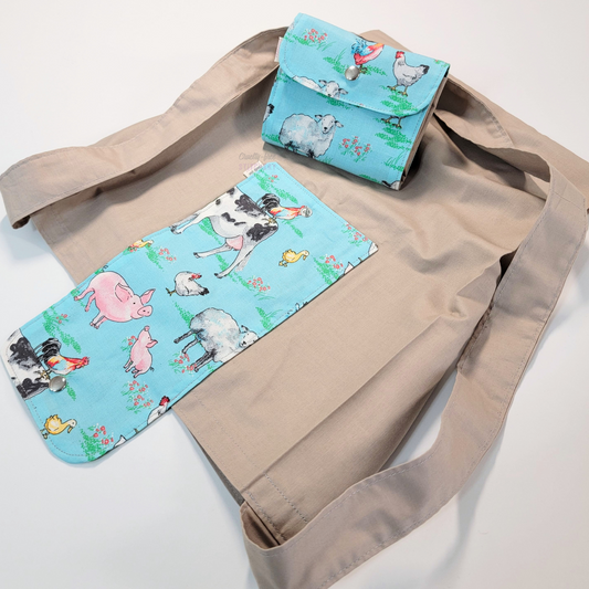 A solid light taupe brown tote bag with a long strap and a light blue with various farm animals printed on the flap - cows, chickens, pigs, ducks. On top of the bag is another bag that has been folded up and snapped together like a wallet.