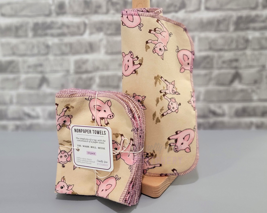 Tan with pink pigs print NonPaper Towels. Shown on a roll, next to a bundled pack. They have rounded corners, stitched with lilac purple thread.