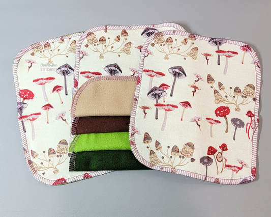 Mushroom print cloth wipes laid out flat, with two on the sides and four in the center with the bottom edges folded up to show the backs are light tan, dark brown, light bud green, or dark green. 