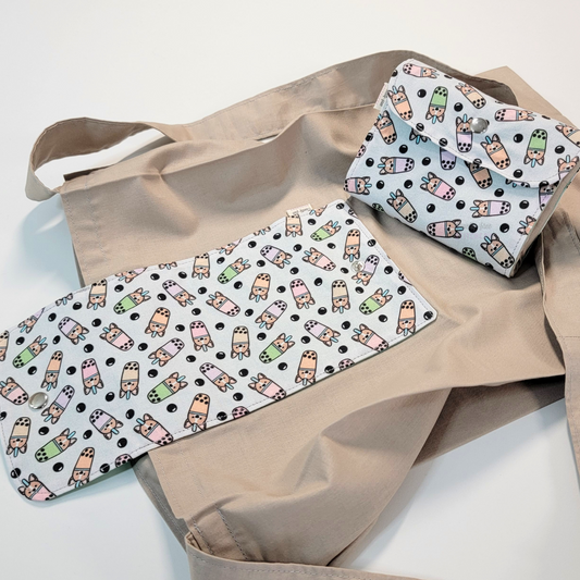A solid light taupe brown tote bag with a long strap and a pale gray flap printed with small boba cups with corgi heads on them. On top of the bag is another bag that has been folded up and snapped together like a wallet.