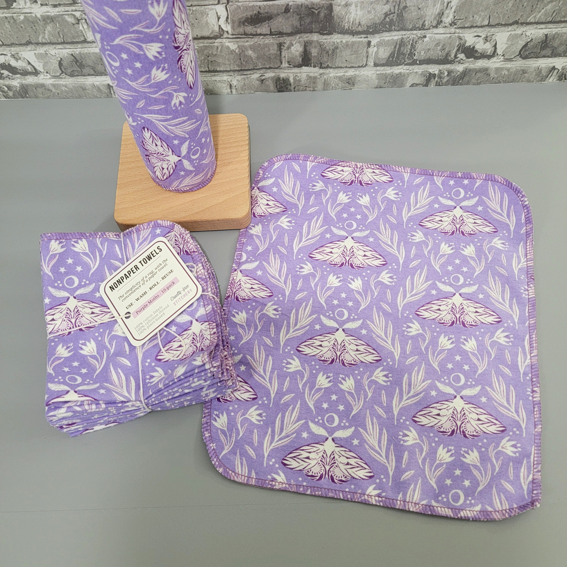 A full purple moths NonPaper Towel laid flat to show the design, next to the bundled pack and the wooden paper towel holder.