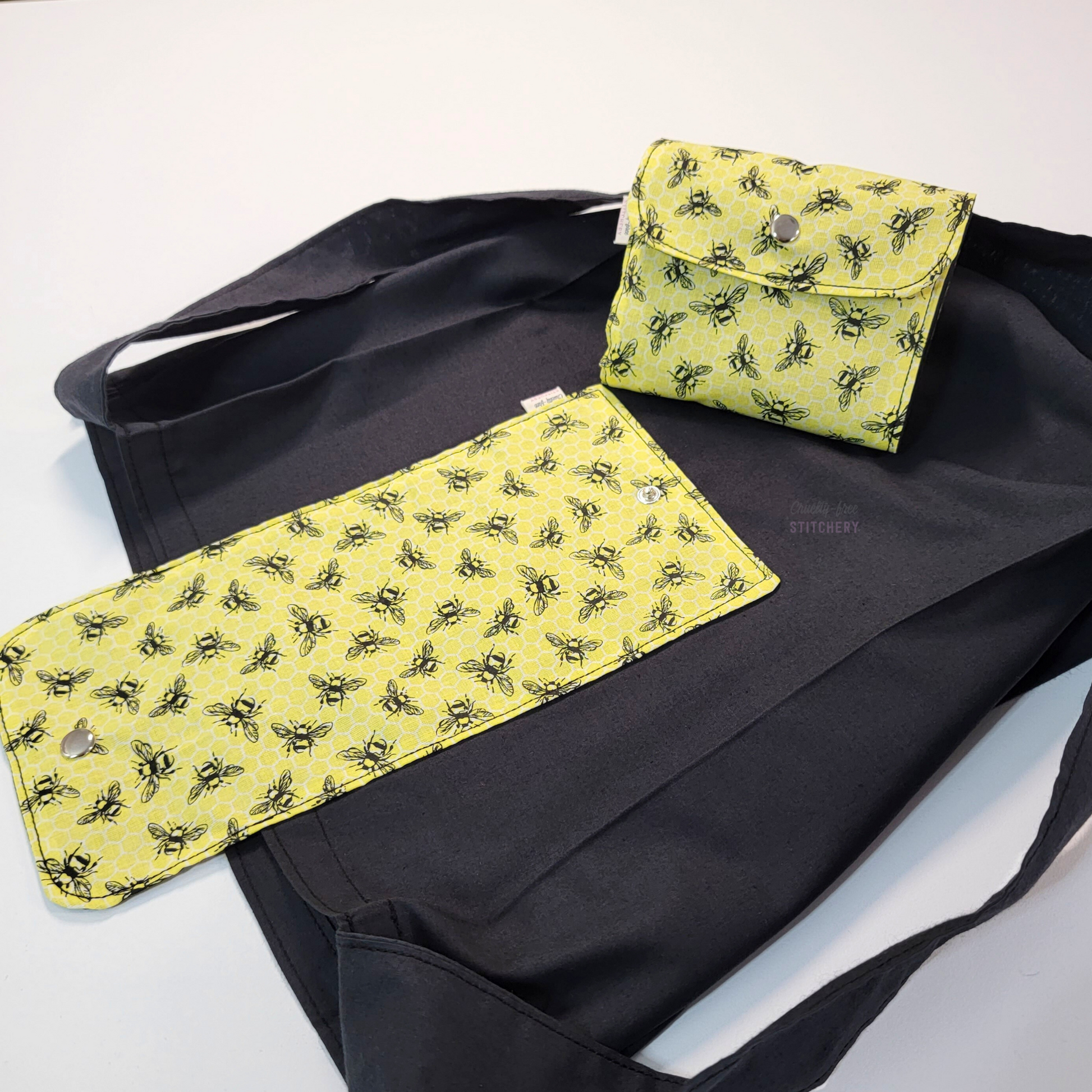 A solid black tote bag with a long strap and a yellow flap printed with small black sketched bees. On top of the bag is another bag that has been folded up and snapped together like a wallet.