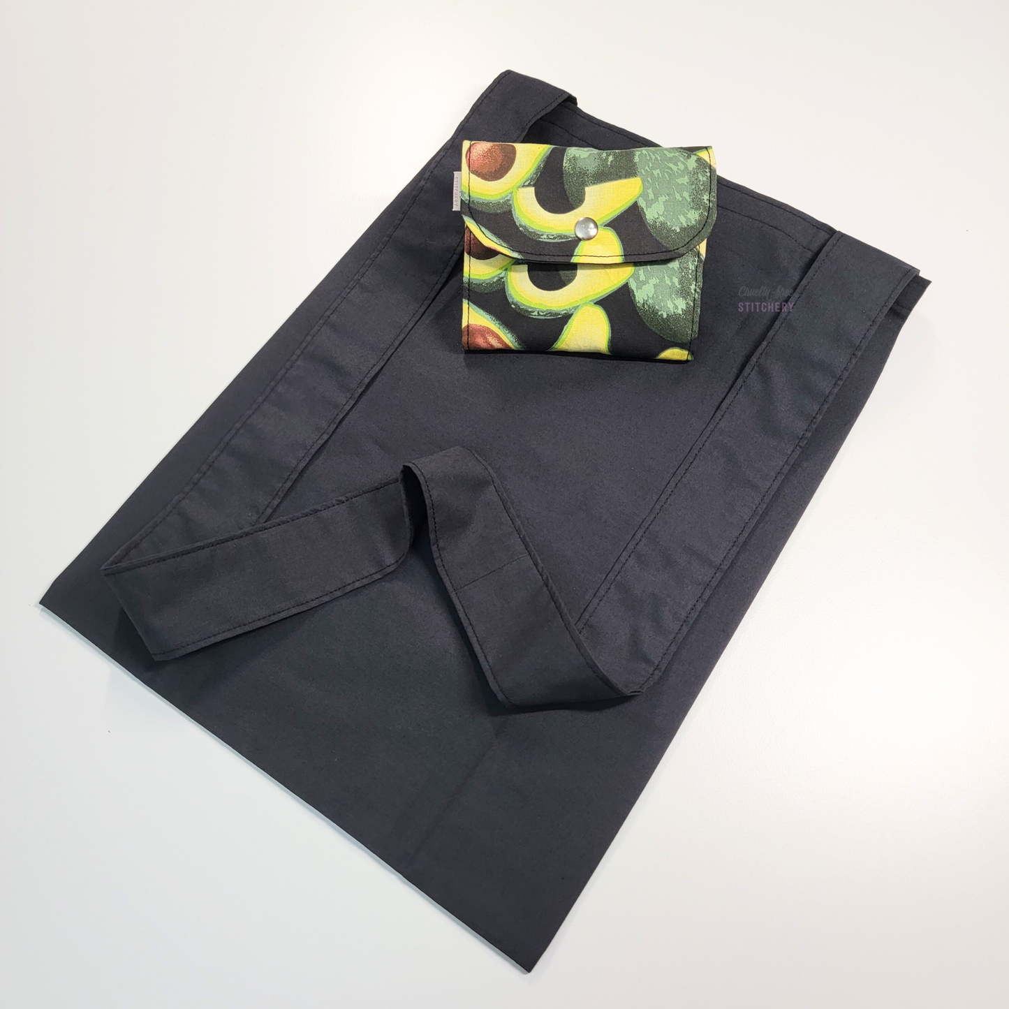 A black tote bag with a long strap. On top of the bag is another bag that has been folded up and snapped together like a wallet. The outer part is a black fabric with avocados.