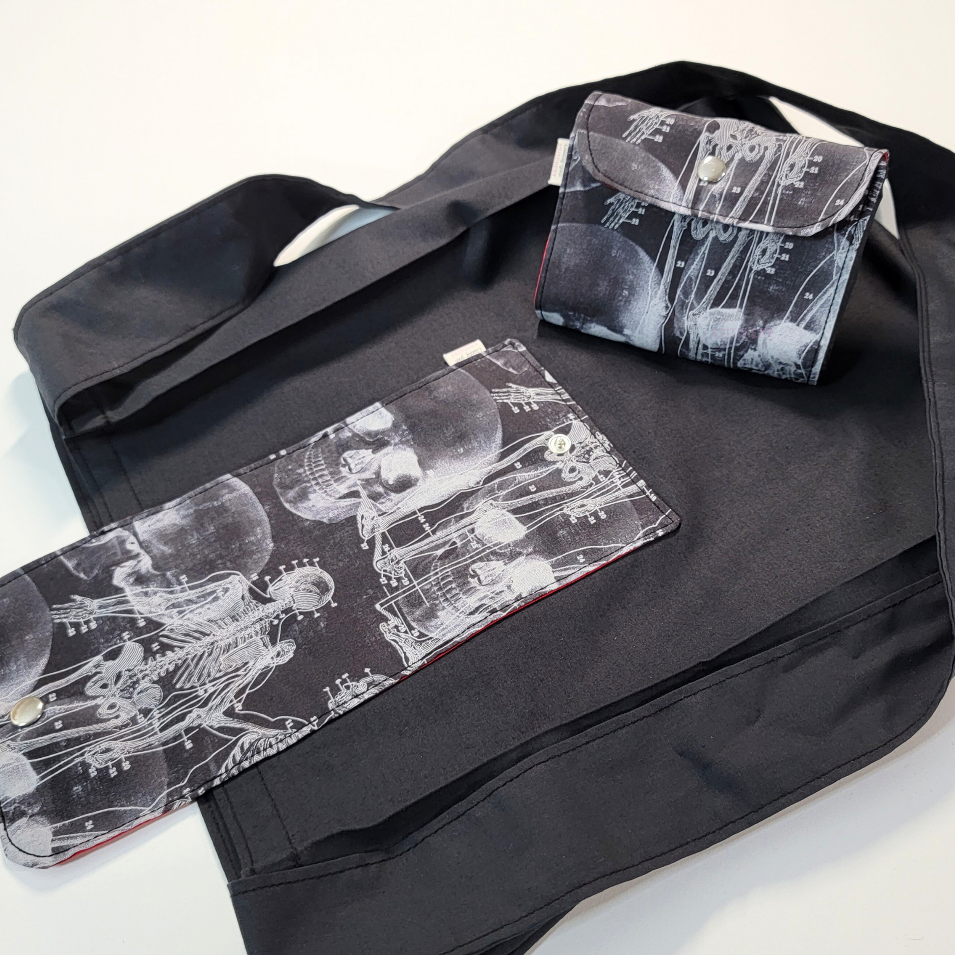 A solid black tote bag with a long strap and a black flap printed with x-ray images of a skull and skeleton. On top of the bag is another bag that has been folded up and snapped together like a wallet.