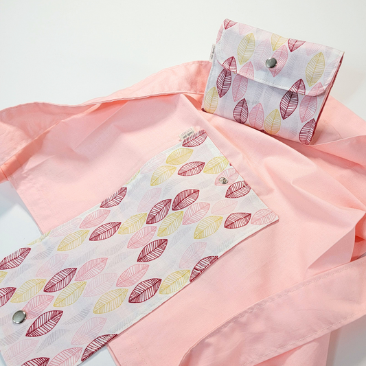 A solid light pink tote bag with a long strap and a white flap printed with sketched leaves in varying shades of pink and yellow. On top of the bag is another bag that has been folded up and snapped together like a wallet.