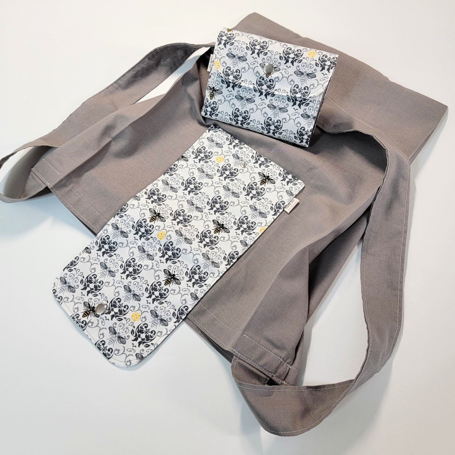 A solid neutral gray tote bag with a long strap and a black and white damask print with small bees and honeycombs on the flap. On top of the bag is another bag that has been folded up and snapped together like a wallet.