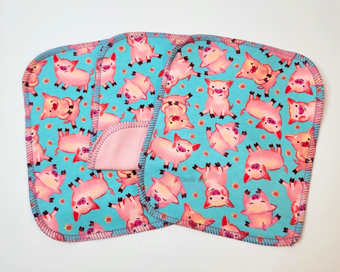 Blue Pigs Reusable Cloth Wipes. Three wipes laid out, with the center one folded back to show the opposite side is light pink. They are a bright aqua blue with small pink pigs and tiny pink flowers. 
