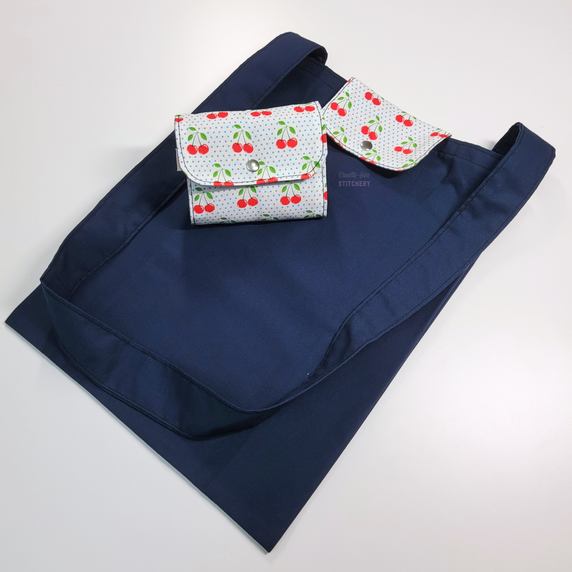 A navy blue tote bag with a long strap and a white flap with cherries and tiny blue polka dots. On top of the bag is another bag that has been folded up and snapped together like a wallet.