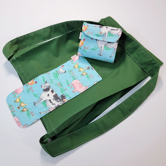 A solid bright leaf green tote bag with a long strap and a light blue with various farm animals printed on the flap - cows, chickens, pigs, ducks. On top of the bag is another bag that has been folded up and snapped together like a wallet.