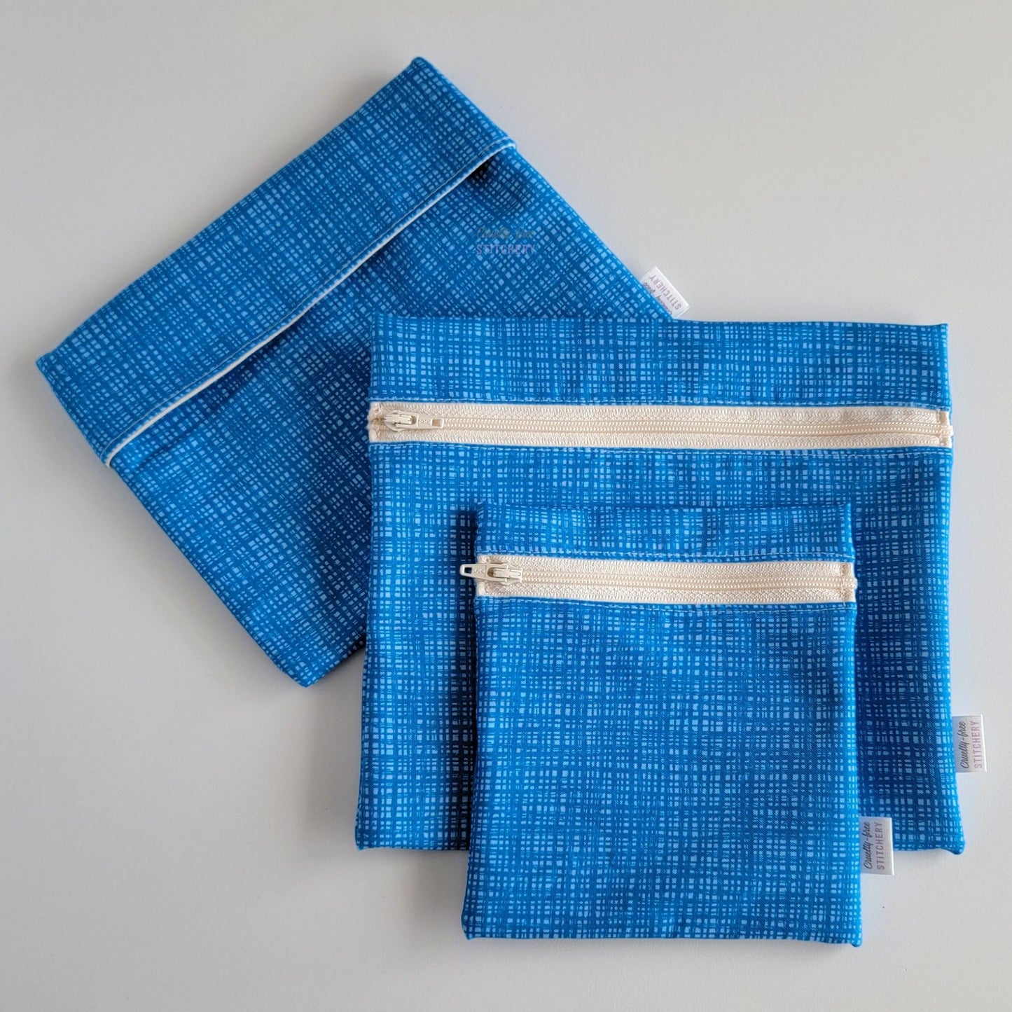 Set of three reusable snack bags with a blue textured print, shown with the foldover style bag closed and behind the zipper styles.