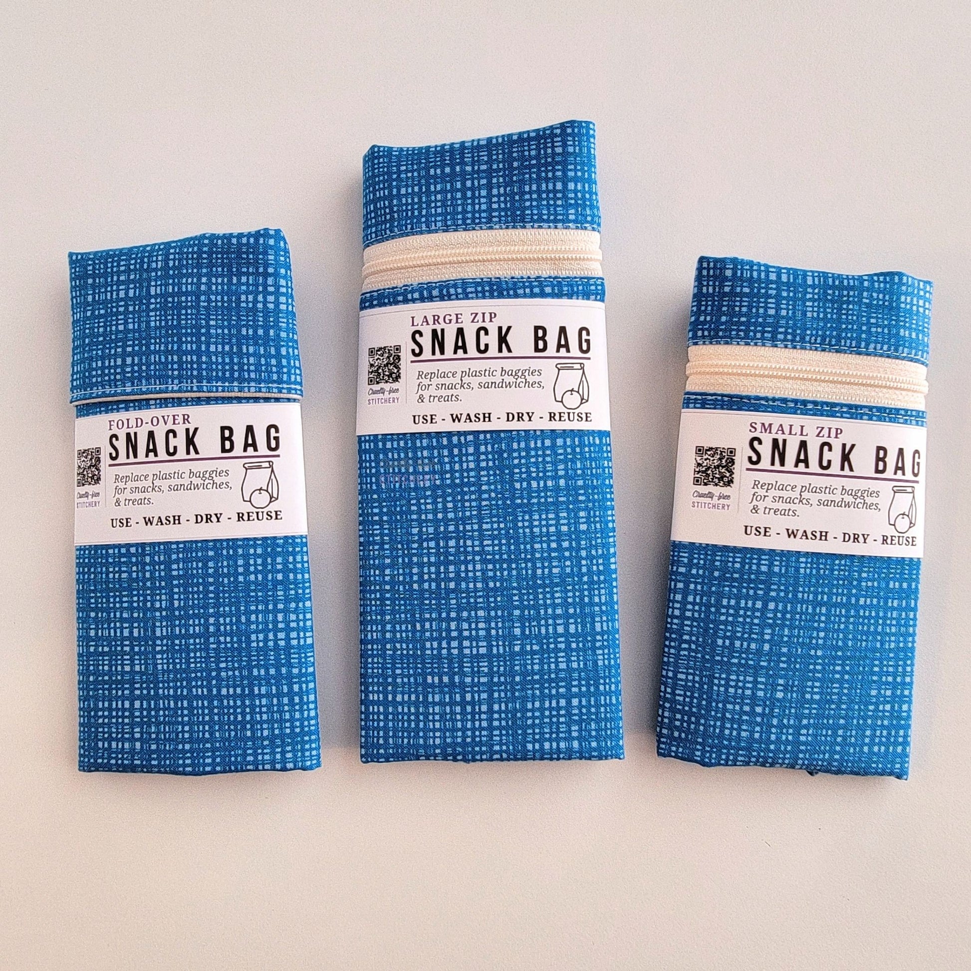 The Blue Texture Snack Bags packaged up. They are folded to be the same width, around 3 inches, and secured with a paper tag wrapped around the middle that says Snack Bag - Replace plastic baggies for snacks, sandwiches, and treats, use, wash, dry, reuse, with an icon of a lunch bag and an apple. 