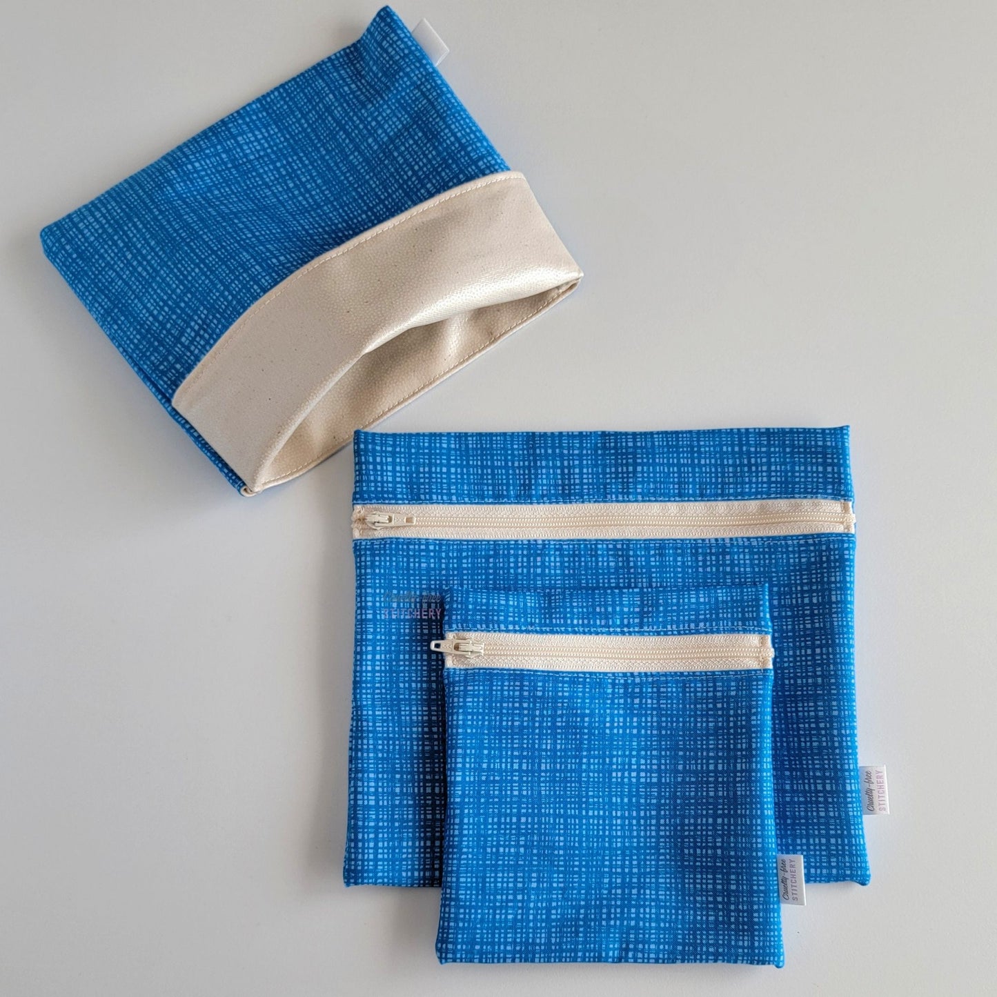 Set of three reusable snack bags with a blue textured print. Two have zippers, one large and one small, and one is a fold-over style. The fold-over one is unfolded to show the beige lining that matches the zippers. The print is a bright blue with a cross-hatch pattern.
