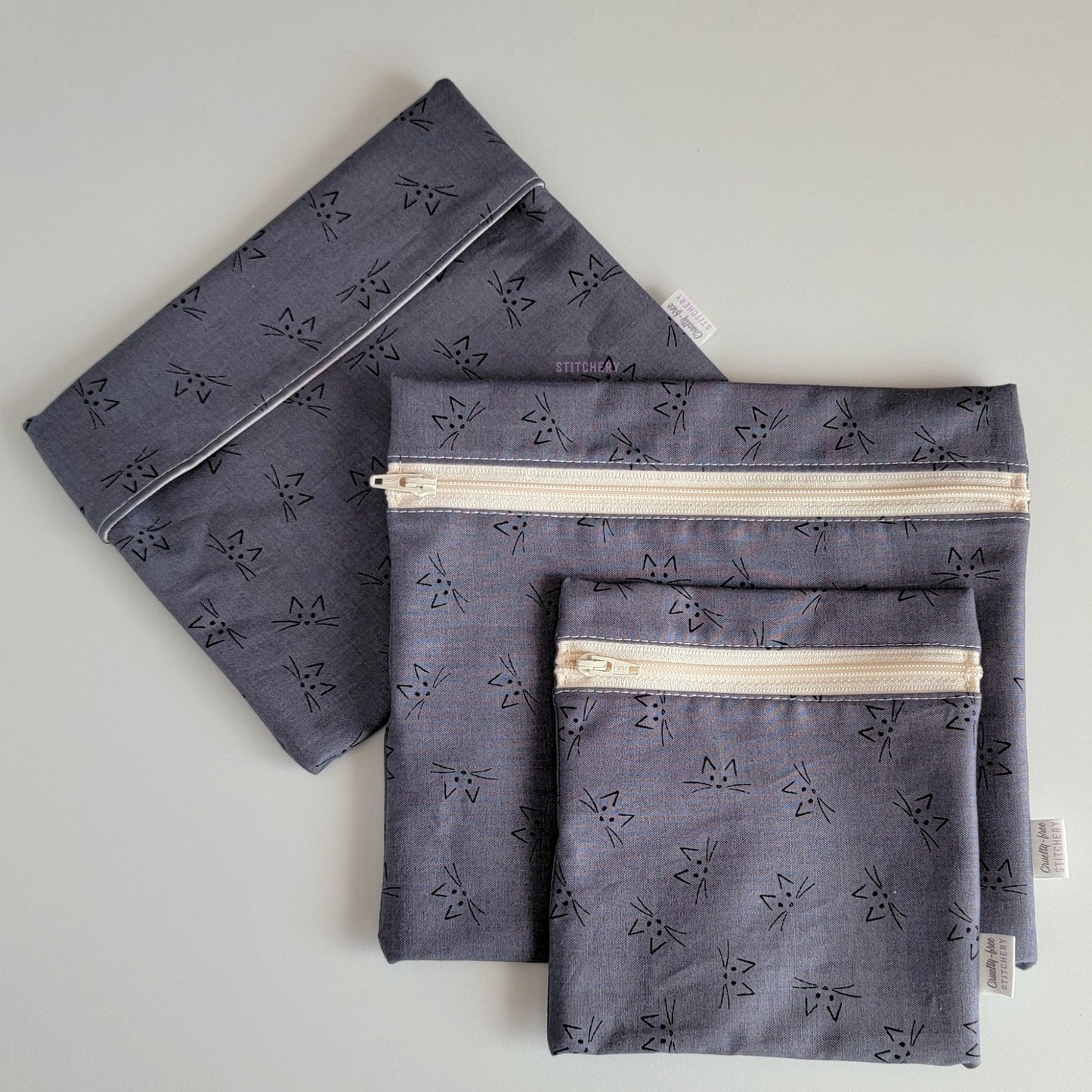 Set of three reusable snack bags with a gray cat faces print, shown with the foldover style bag closed and behind the zipper styles. 