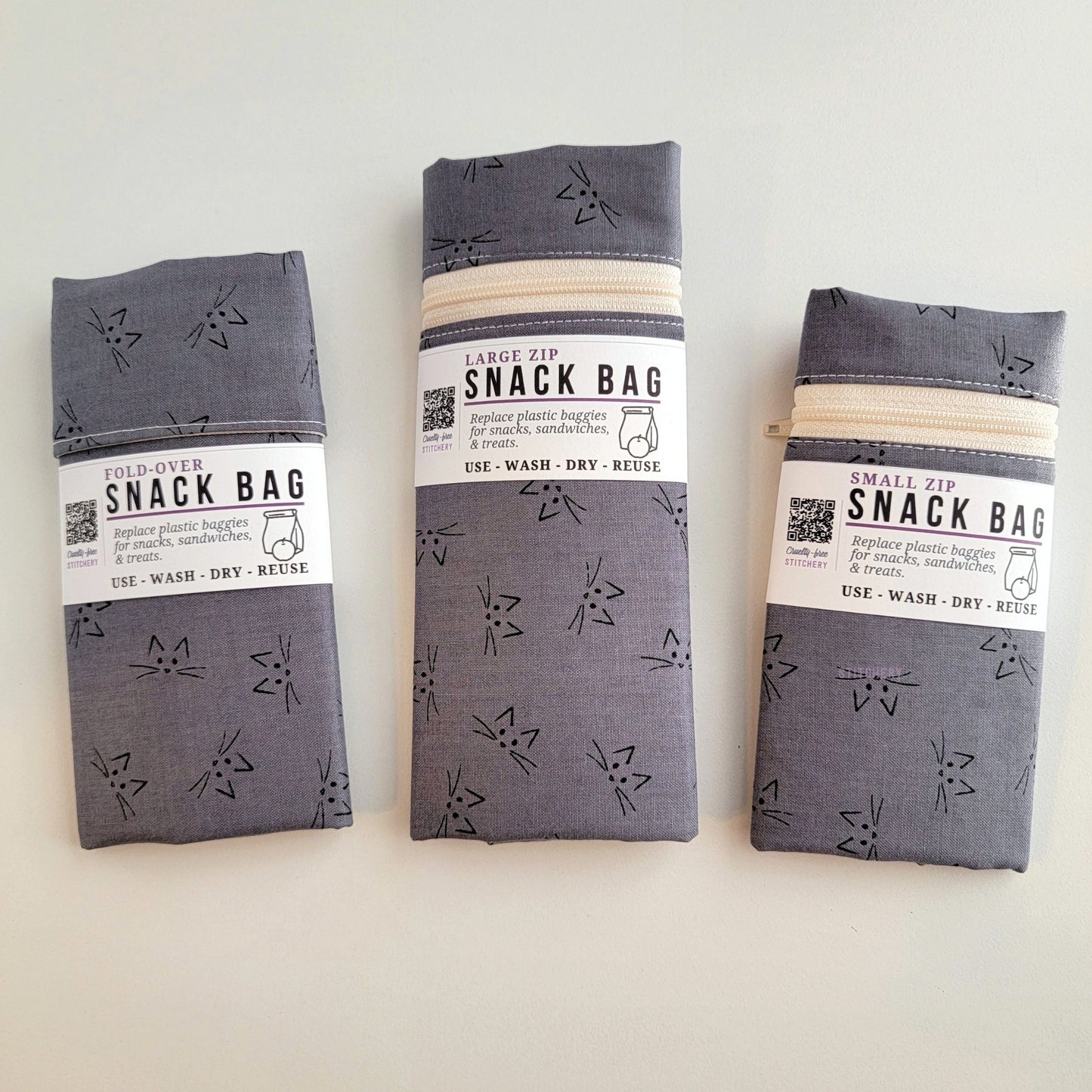 The Gray Cat Faces Snack Bags packaged up. They are folded to be the same width, around 3 inches, and secured with a paper tag wrapped around the middle that says Snack Bag - Replace plastic baggies for snacks, sandwiches, and treats, use, wash, dry, reuse, with an icon of a lunch bag and an apple.